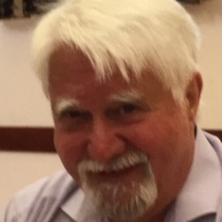 An older man with white hair, a mustache, and beard, smiling while wearing a light purple shirt indoors.