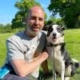 A man sits on grass beside a black and white dog on a sunny day in a park with green trees.