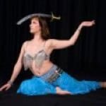 A woman in a belly dance costume balances a sword on her head while posing on the floor against a black background.