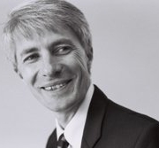 Black and white photo of a smiling man with light hair, wearing a suit and tie, looking to the side.