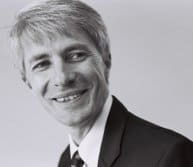 Black and white photo of a smiling man with light hair, wearing a suit and tie, looking to the side.